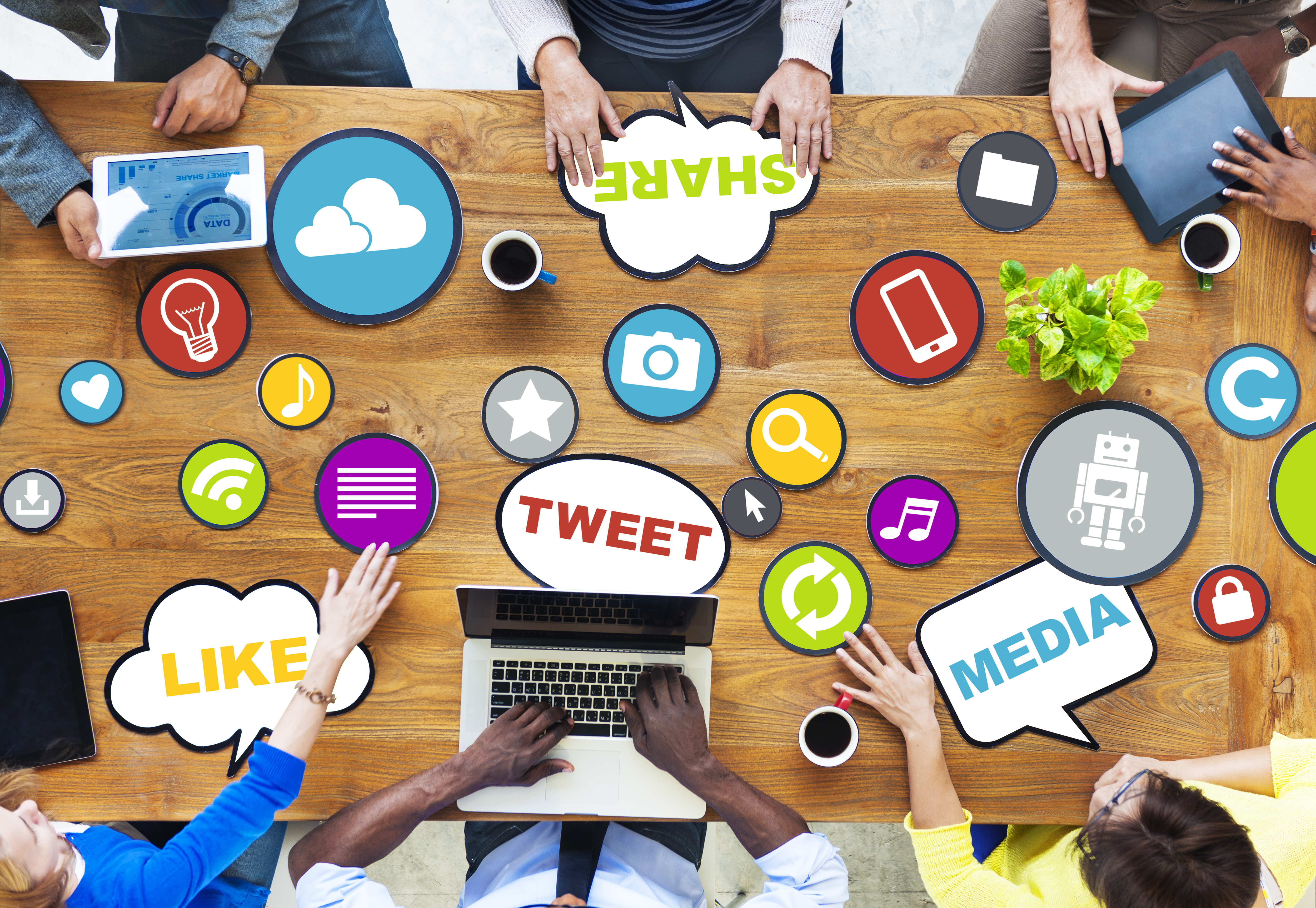 Aerial view of multiple people sitting at a table with latops and tablets. The words 'like, share, tweet, and media' surround them on the table.